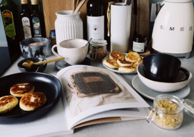 One portion of homemade cheese pancakes in frying pan and another on plate locating near opened stylish magazine among kitchen utensils and appliances in bright kitchen at home
